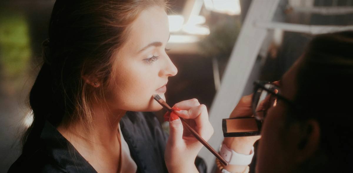 A close-up photo showing hands applying blush and highlighter on a model's cheekbones.